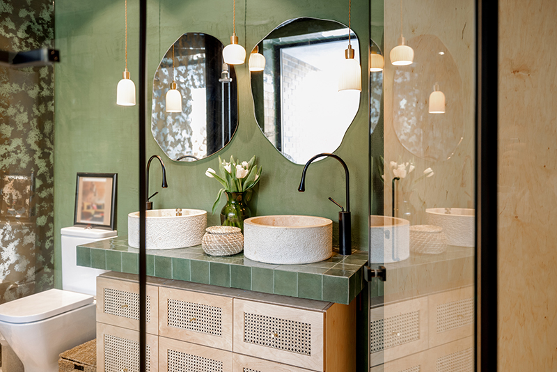 Bathroom interior with stone wash basins, modern faucets, irregularly shaped mirrors and wooden rattan commode in green and beige tones in natural boho style