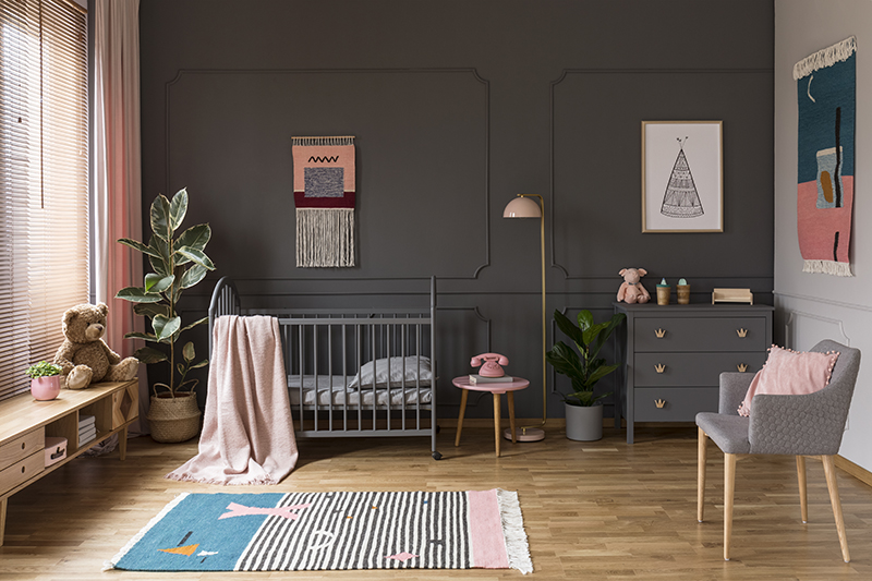 Real photo of a grey crib standing next to a pink stool, a lamp and cupboard in grey baby room interior also with armchair, rug and poster